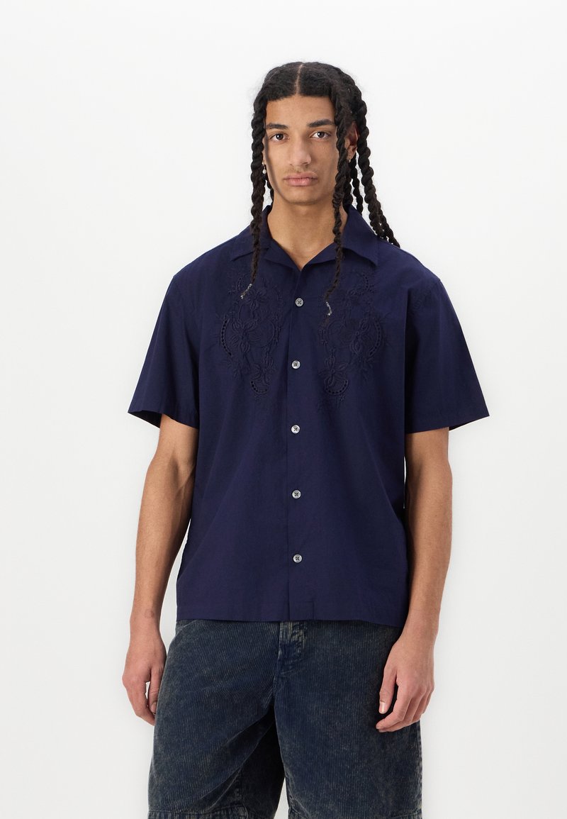 Camisa de manga corta azul marino con detalles bordados en el frente, cierre de botones y un color clásico. Se exhiben pantalones cortos de mezclilla de Malibu.