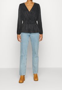 Top nero a maniche lunghe con scollatura a V profonda, tessuto plissettato e un leggero luccichio. Abbinato a jeans azzurri e stivali platform beige.