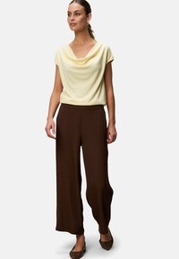 Gelbes Top mit Wasserfallkragen und kurzen Ärmeln, kombiniert mit lockeren, braunen Hosen. Glatter Stoff; leichtes Material; klassisches Design. Braune Schuhe runden das Outfit ab.