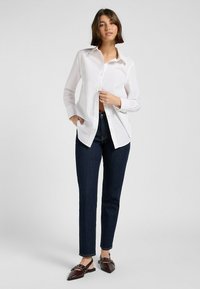 Witte langarmpolo met button-down kraag, open gedragen over donkere spijkerbroeken en bruine schoenen met decoratieve hardware en puntige neuzen.