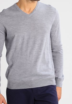 Mand iført en lysegrå V-hals langærmet sweater og mørkeblå bukser mod en ensfarvet baggrund.