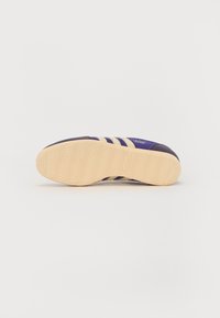 Sneaker violette avec trois rayures beiges, semelle en caoutchouc présentant un motif en chevrons et détail du logo près du talon.