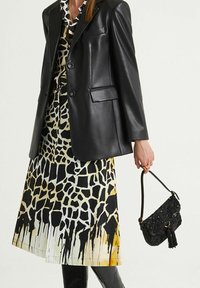 Blazer in pelle nera indossato su un vestito a fantasia con stampa di giraffa e sfumature dal giallo al nero. Piccola borsa nera con perline e nappina.