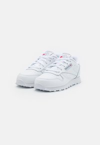 Baskets blanches avec un cuir lisse sur le dessus, une languette rembourrée et une semelle en caoutchouc texturée. Comprend des lacets et des accents de logo sur le côté.