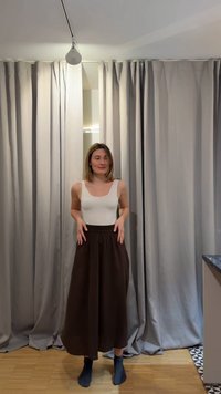 Γυναίκα με μαλλιά μέχρι τον ώμο που φοράει ένα λευκό αμάνικο τοπ, φαρδιά καφέ παντελόνια και σκούρες κάλτσες, στέκεται μπροστά από μπεζ κουρτίνες.