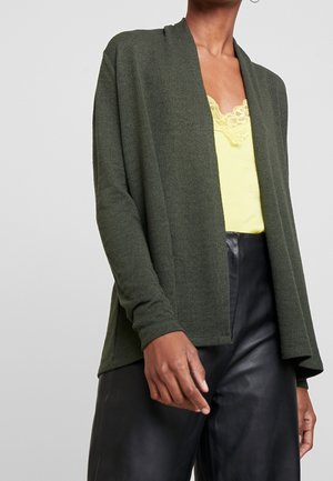 Personne portant un cardigan vert foncé ouvert sur un haut jaune orné de dentelle et un pantalon en cuir noir, sur un fond uni.