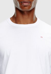 Hvid kortærmet t-shirt med rund hals og et lille rødt logo på venstre brystområde, lavet af glat stof med et enkelt design.