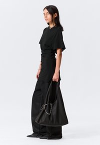 T-shirt preto ajustado com mangas curtas, combinado com calças largas pretas. O modelo porta uma mala de couro preta com detalhes em corrente prateada.