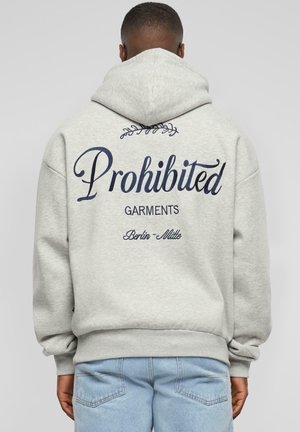 Homme portant un sweat à capuche gris clair avec le texte "Prohibited Garments Berlin-Mitte" dans le dos, associé à un jean bleu clair.