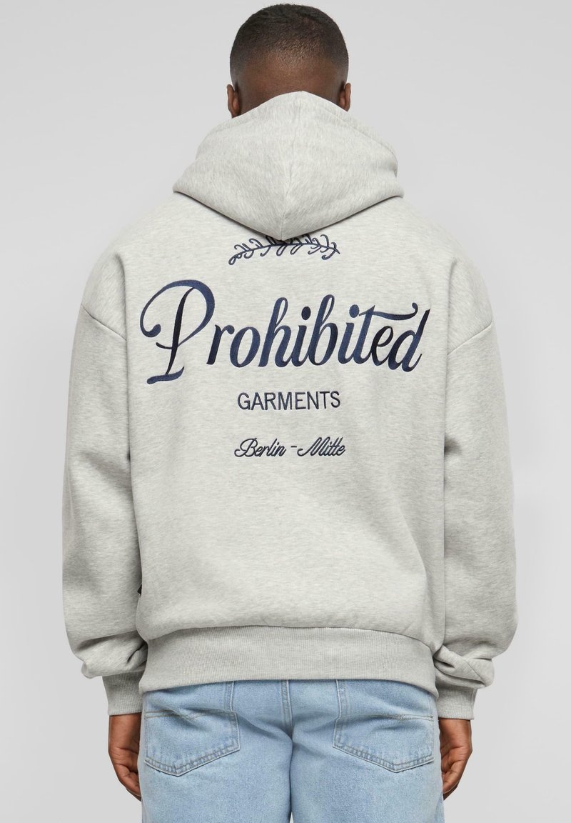 Homme portant un sweat à capuche gris clair avec le texte "Prohibited Garments Berlin-Mitte" dans le dos, associé à un jean bleu clair.