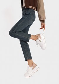 Sneakers bianchi con un accento beige testurizzato sul tallone, lacci bianchi e una suola spessa. Indossati con jeans blu scuro alla caviglia.