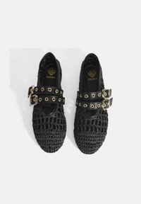 Schwarze gewebte Schuhe mit strukturiertem Design, ausgestattet mit doppelten Riemenakzenten und goldenen Beschlägen. Die Innensohle zeigt den Markennamen "DESA."