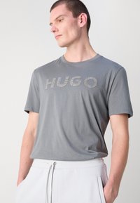 Pelēka kokvilnas T-kreklu ar nedaudz pieguļošu dizainu. "HUGO" logo ar teksturētu metāla druku krūšu daļā. Īsas piedurknes, apaļš kakla izgriezums.