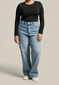 Osoba ubrana w czarną bluzkę z długim rękawem, jasnoniebieskie szerokie spodnie typu dzwony i białe trampki, stojąca na tle jednolitego tła.