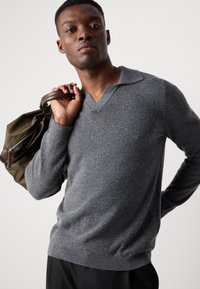 Gray wool pullover with a V-neck and ribbed cuffs, worn by a person holding a dark green bag with brown leather accents.