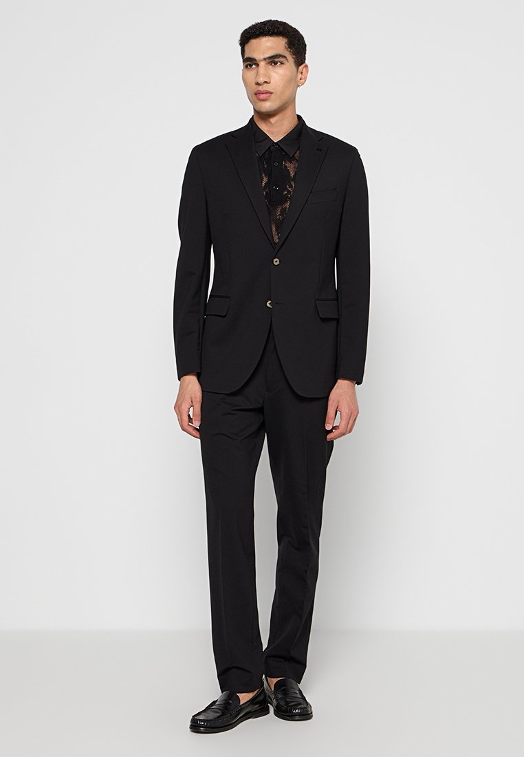 Un giovane uomo sta in piedi con sicurezza in un completo nero aderente e una camicia, mostrando un look moderno e alla moda su uno sfondo semplice.