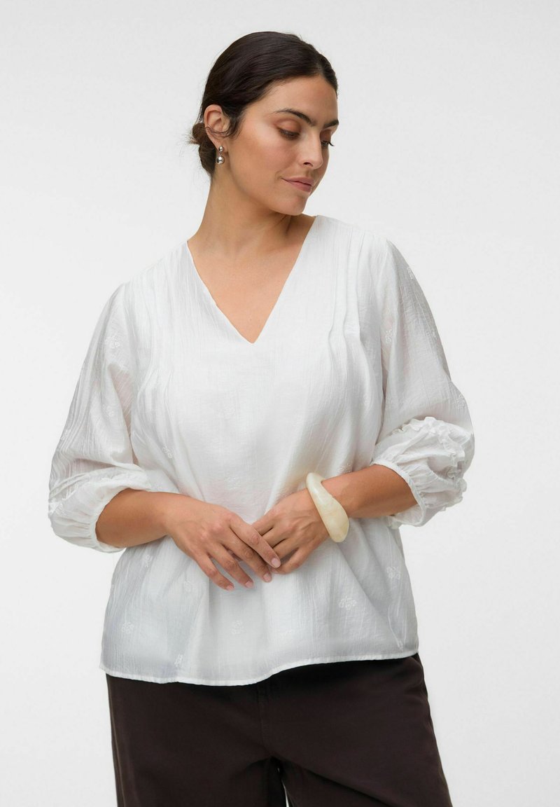 Femme aux cheveux foncés portant un chemisier blanc texturé à manches trois-quarts et un pantalon foncé, regardant vers le bas, les mains jointes devant.