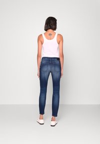 Mujer de pie de espaldas, vestida con una camiseta sin mangas blanca, jeans skinny azules, zapatos rosa claro, con un tatuaje floral en la parte superior de la espalda.