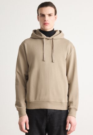 Beige hoodie med snor og et lille logo på brystet. Har en afslappet pasform og ribbede manchetter, og er parret med sorte jeans.