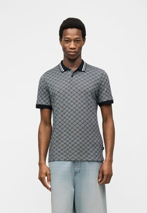 Jeune homme debout, portant un polo gris à motifs avec col et poignets noirs, et un jean bleu clair, sur fond blanc.