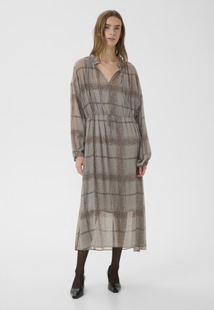 Frau steht und trägt ein langes, lockeres graues Kleid mit braunem Karomuster, langen Ärmeln, schwarzen Strumpfhosen und schwarzen Schuhen mit niedrigem Absatz.