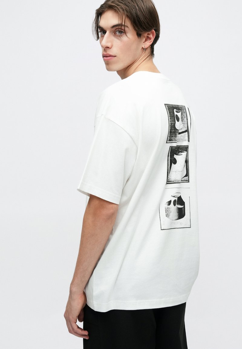 Camiseta de algodón blanca con un corte relajado que presenta tres impresiones gráficas negras en la parte posterior. Mangas cortas y cuello redondo.