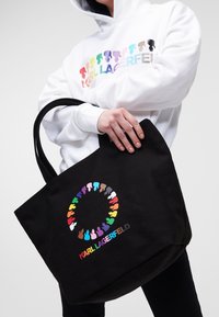 Černá plátěná taška s kulatým vícerozměrným siluetovým designem a logem v živých písmenech. Dvě pevné uši pro nošení.