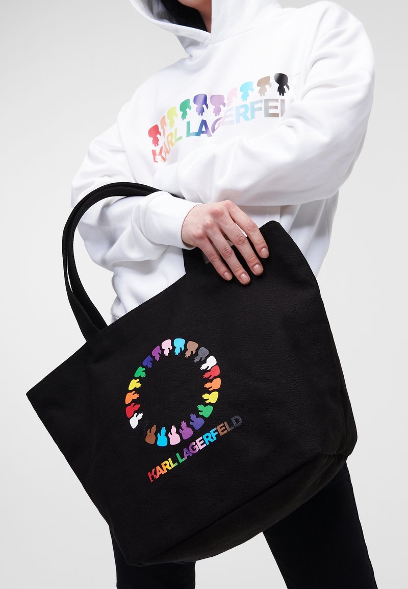 Černá plátěná taška s kulatým vícerozměrným siluetovým designem a logem v živých písmenech. Dvě pevné uši pro nošení.