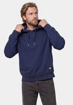 Mand med skæg justerer hætten på en mørkeblå sweatshirt, iført mørke jeans, kigger til siden mod en ensartet lys baggrund.