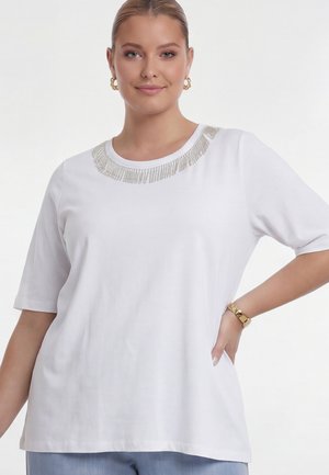 Woman wearing white short-sleeve top with silver decorative fringe along neckline, gold hoop earrings, and gold watch on left wrist.