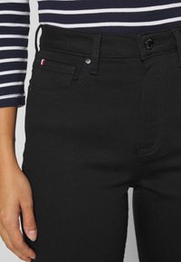 Jeans de mezclilla negra con un diseño ajustado, que incluyen un bolsillo frontal, herrajes plateados y un pequeño acento de logo tricolor en el costado.