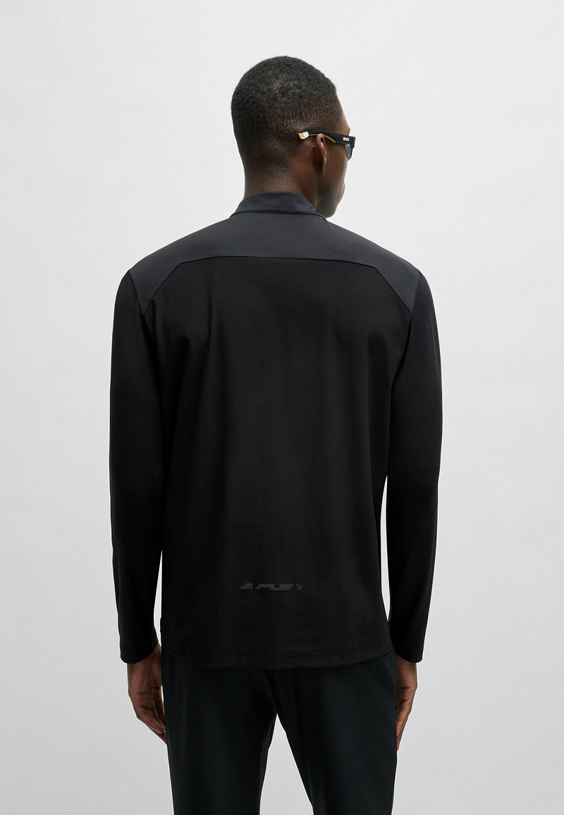 Camisa negra de manga larga con una textura suave y tela mate contrastante en los hombros. Detalle de logo sutil en la cintura trasera.