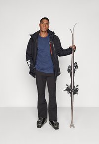 Skisportutstyr viser en mann i en svart vanntett jakke med røde detaljer, marineblå skjorte, svarte bukser og svarte skistøvler, som holder ski.
