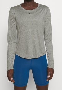 Šedé sportovní tričko s dlouhým rukávem a kulatým výstřihem s černým logem, doplněné modrými kompresními šortkami, hladká textura, přiléhavý střih.