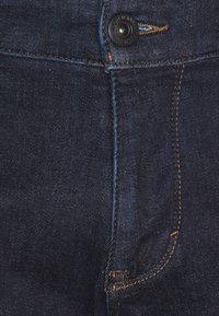 Mörkblå denimjeans med knäppning, kopparstickning och en slät yta med en svag glans. Inga synliga fickor.