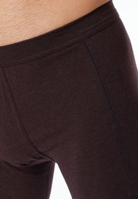 Nahaufnahme einer Person, die dunkelbraune, schmal geschnittene Hosen trägt, mit sichtbaren Nähten und leichtem Körperhaar über dem Bund.