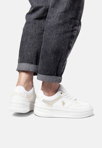 Bílé tenisky vyrobené z umělého materiálu s perforovanými detaily a zlatým logem. Zaoblená špička, tkaničkový design a silná podrážka.