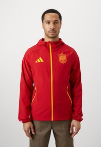Giacca sportiva rossa con dettagli gialli. Presenta una chiusura a zip, cappuccio e logo. La giacca ha un design elegante ed è realizzata in materiale leggero.