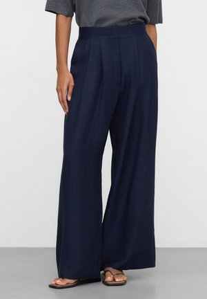 Persoon draagt marineblauwe wijde broek, grijs kort mouwen top en zwarte teenslippers, staand tegen een effen witte achtergrond.