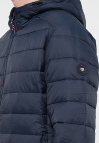 Giubbotto imbottito blu navy con cappuccio, caratterizzato da cuciture orizzontali, chiusura con zip e una patch circolare del marchio sulla manica sinistra.