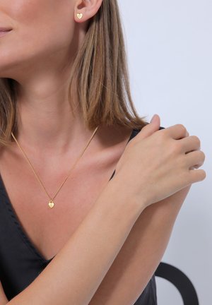 Boucles d'oreilles en forme de cœur en or et un collier assorti sur une épaule nue, avec une chaîne tordue et un petit pendentif en forme de cœur sur un haut sombre.