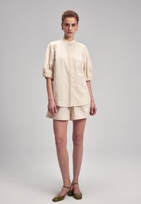 Cremfarbene Bluse mit strukturiertem Muster und Puffärmeln, kombiniert mit passender Shorts. Das Modell trägt grüne Ballerinas mit Schnallen-Details.