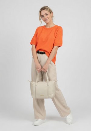 Lachende vrouw in een oranje top met korte mouwen en beige broek, die een beige handtas vasthoudt en met één gebogen been staat tegen een effen achtergrond.