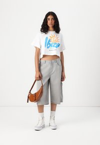 Λευκό cropped t-shirt με γραφικό "Ibiza Sun" σε μπλε και πορτοκαλί χρώμα, συνδ�υασμένο με γκρι τζιν culottes και καφέ υφασμάτινη τσάντα. Λευκά αθλητικά παπούτσια.