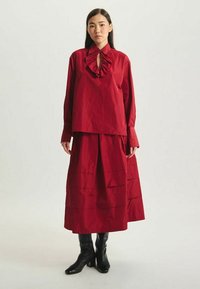 Haut rouge à col froncé, associé à une jupe longue assortie avec des plis horizontaux. Porté avec des bottes noires jusqu'aux genoux.