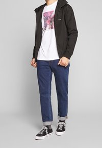 Chaqueta negra con cremallera y capucha, camiseta gráfica blanca, vaqueros azules remangados y zapatillas negras con suelas blancas. Conjunto casual.