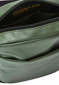 Interior de bolsa sintética verde con cierre de cremallera. Incluye una etiqueta de logo, textura suave y un bolsillo plano en el interior.