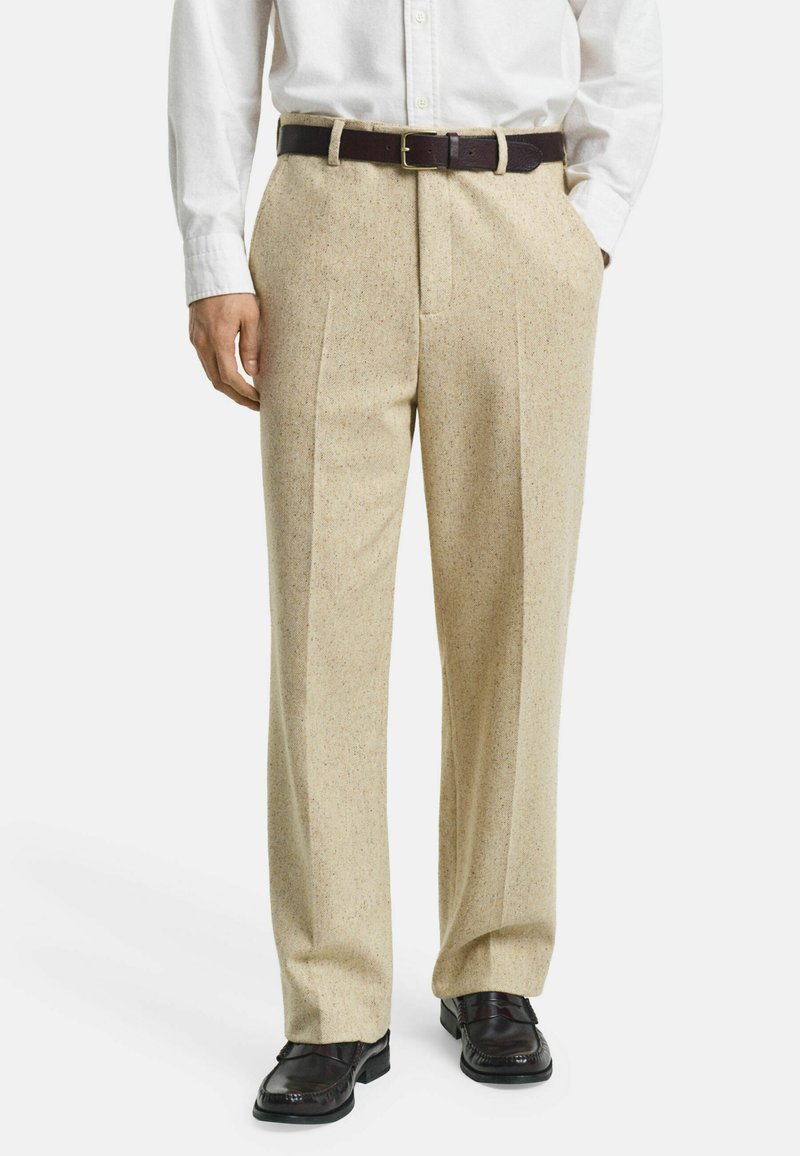 Pantalons beiges sur mesure en mélange de laine texturée, avec des plis à l'avant et une ceinture en cuir marron. Associés à des chaussures en cuir noires.