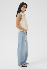 Béžová bezrukávová košeľa s golierom, skombinovaná so svetlo modrými širokými džínsami s rozstrapkaným lemom. Čierne ploché sandále dopĺňajú vzhľad.