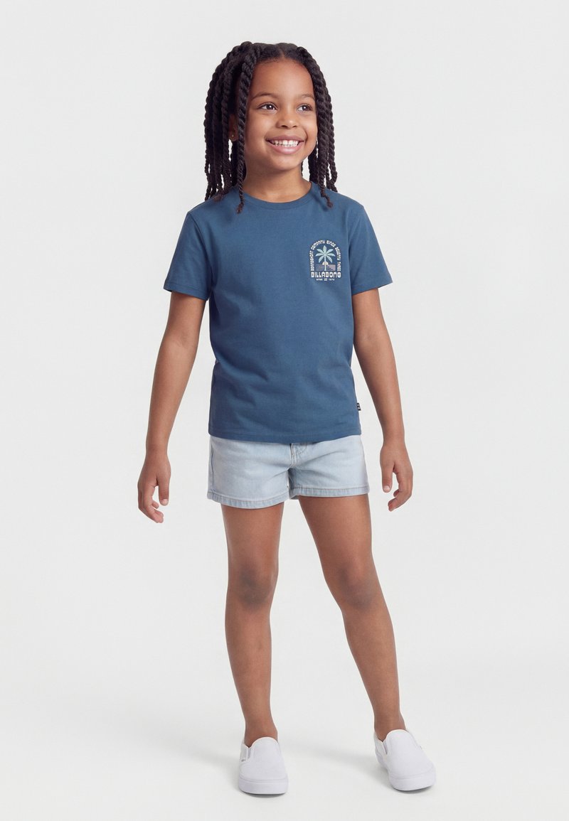 Lachend meisje met ingevlochten haar, een blauw T-shirt met opdruk, licht denim short en witte instapschoenen, staand tegen een effen achtergrond.